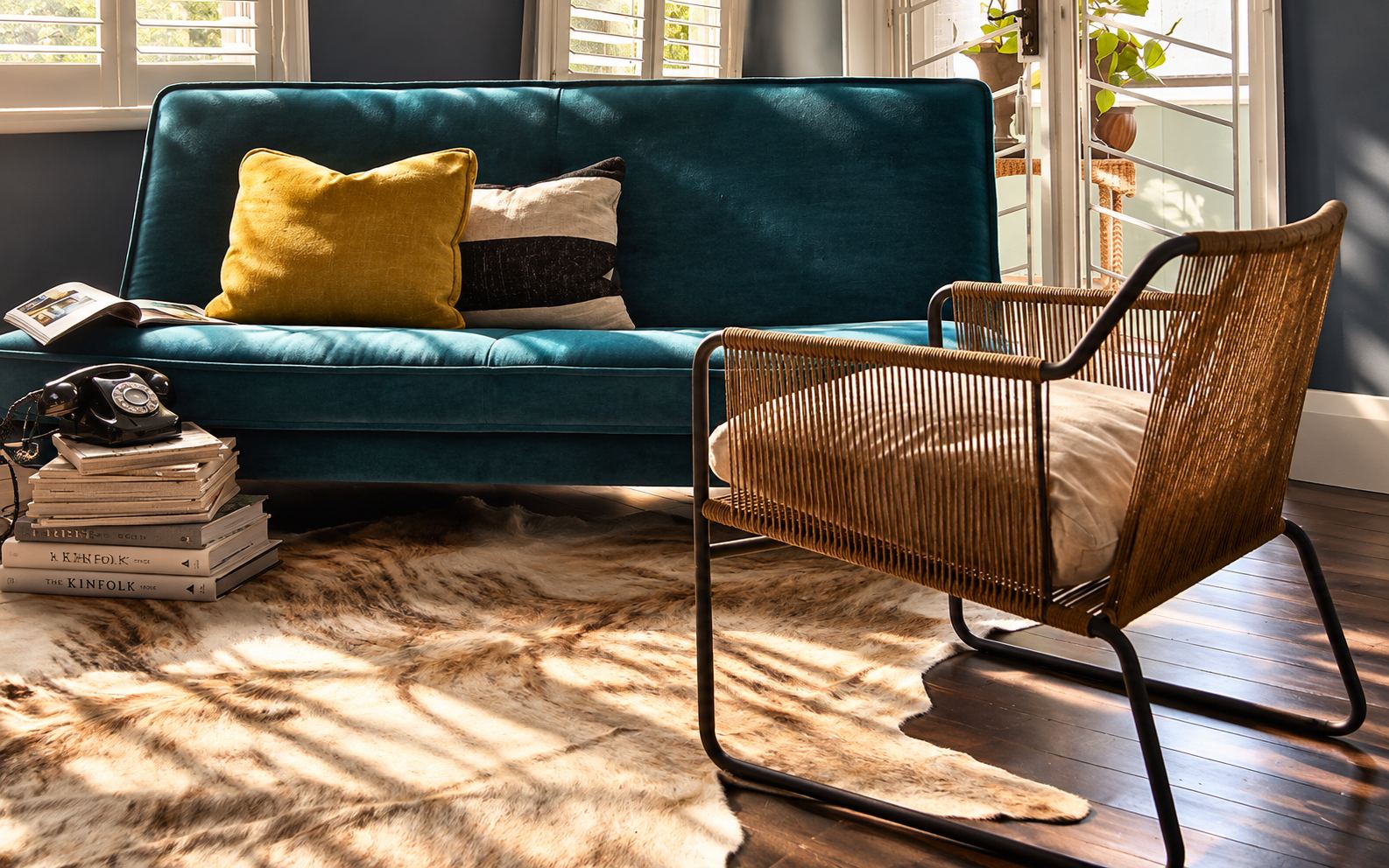 Teal sofa and rope armchair on a cowhide rug in afternoon light, with books stacked beside the sofa